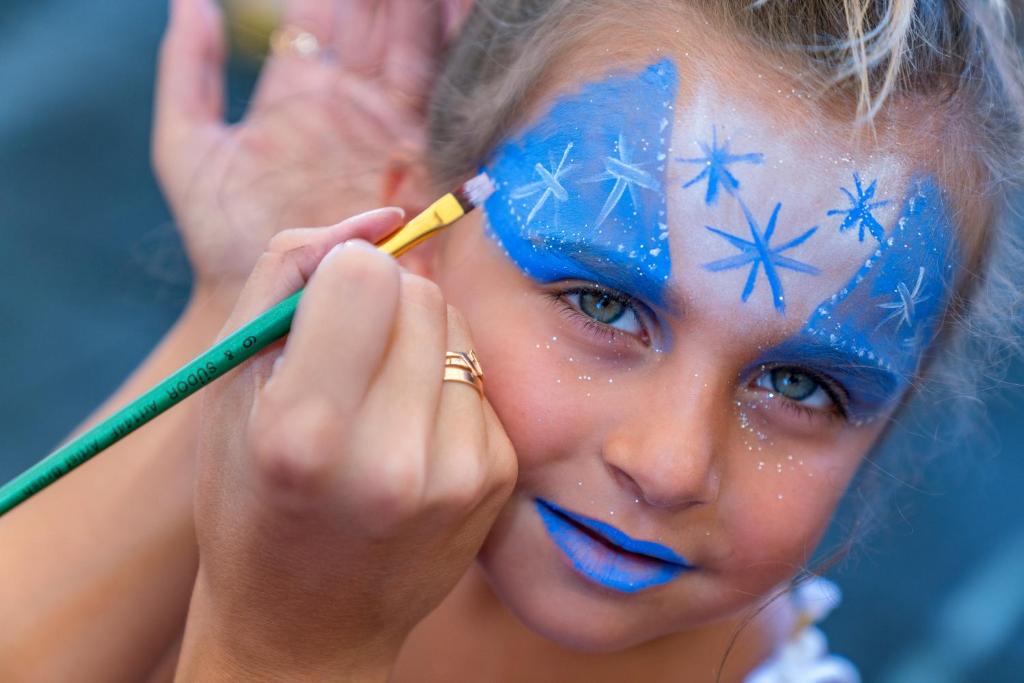 Bērns ar zilu sejas krāsu un sniegpārslām uz pieres saņem sejas krāsošanu. Fons ir neskaidrs, izceļot bērna izteiksmīgās acis un radošo mākslas procesu.