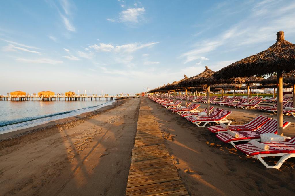 Pludmales skats ar saulainu debesu fonu, kur gar krastu redzami saulessargi un pludmales krēsli. Krasta koka tilts vijas gar smilšaino pludmali, radot relaksējošu atmosfēru.