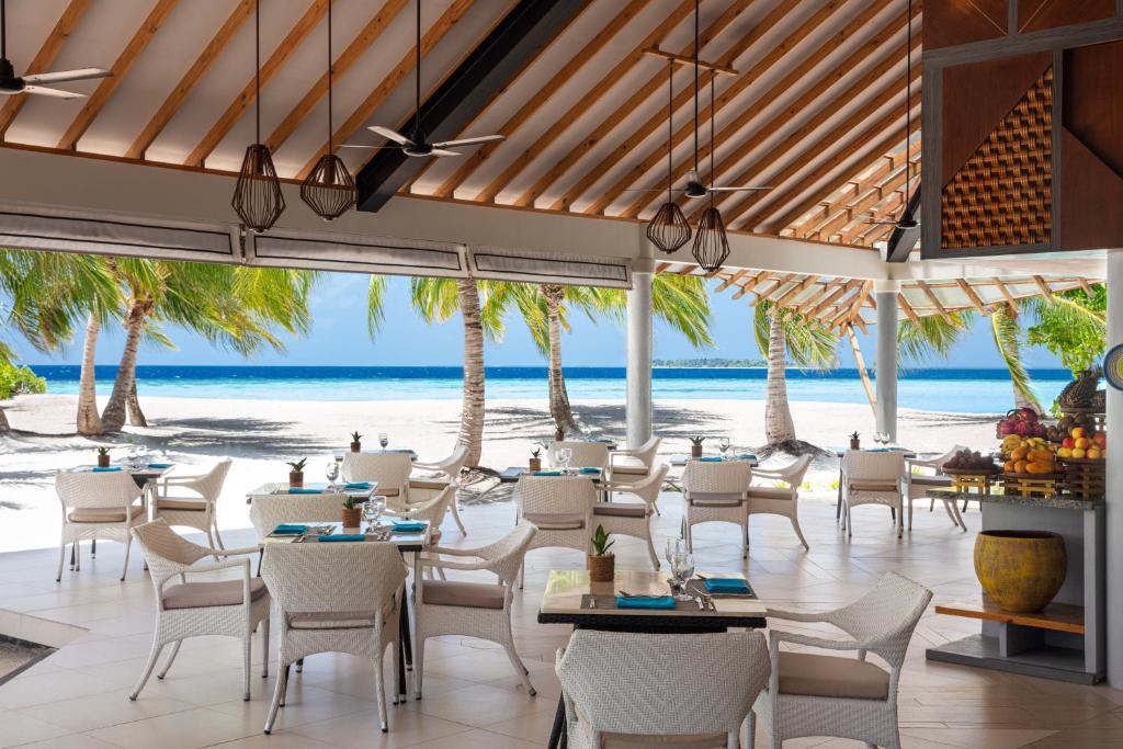 Moderns restorāns ar skatu uz pludmali un palmām, kurā ir eleganti galdi un krēsli. Telpā ir daudz gaismas un atvērta arhitektūra, radot relaksējošu atmosfēru.