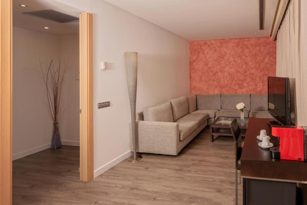 Mūsdienīgs viesnīcas numurs ar pelēku dīvānu, televizoru un nelielu kafijas stūrīti. Sienas ir dekorētas ar siltu sarkano toni, radot mājīgu atmosfēru.