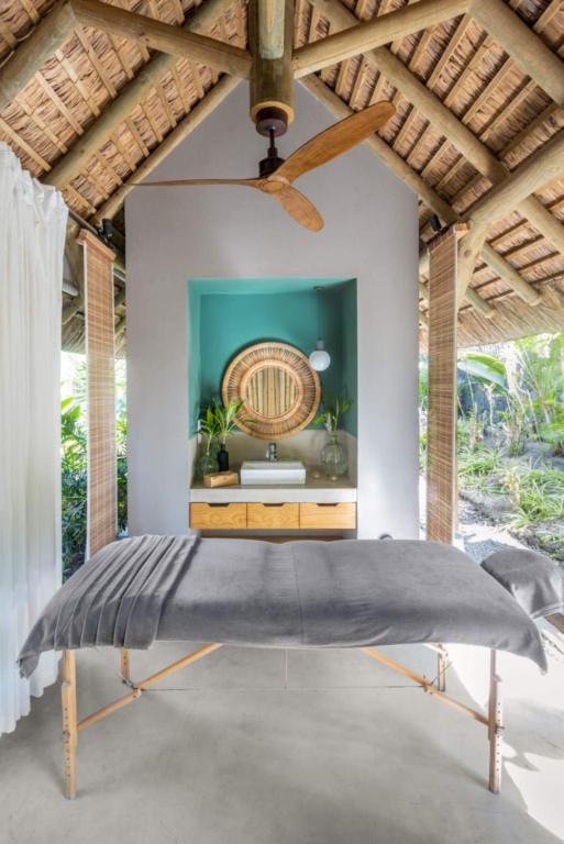 Relaksējoša masāžas telpa ar dabīgu apgaismojumu, koka griestiem un modernu interjeru. Galda virsma ir pārklāta ar pelēku audumu, radot mierīgu atmosfēru.