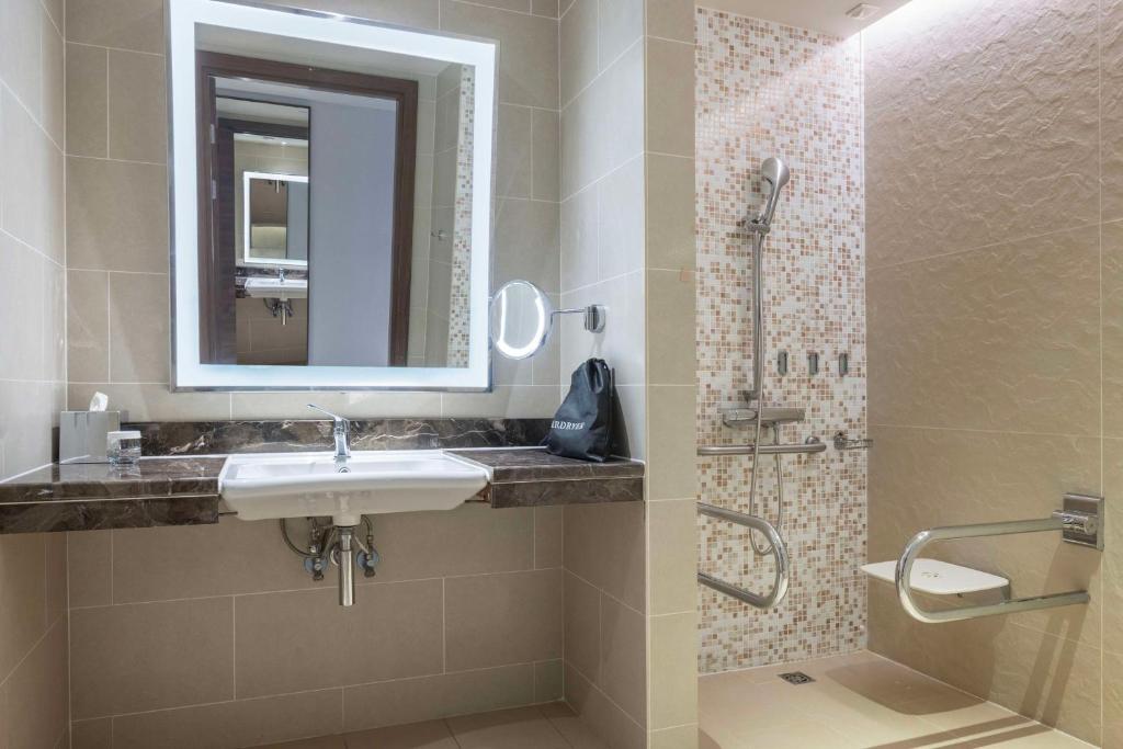Mūsdienīgs vannas istabas interjers ar elegantu spoguli un marmora izlietni, apvienojot modernu dizainu ar funkcionālām detaļām. Dušas zona ir aprīkota ar rokas dušu un drošības barjerām.