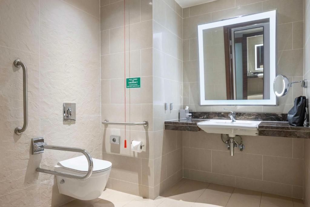 Mūsdienīga vannas istaba ar pieejamības elementiem, tostarp rokturiem un lielu spoguli. Telpa ir gaiša un eleganta, ar akmens izskata virsmām un sanitārajiem izstrādājumiem.