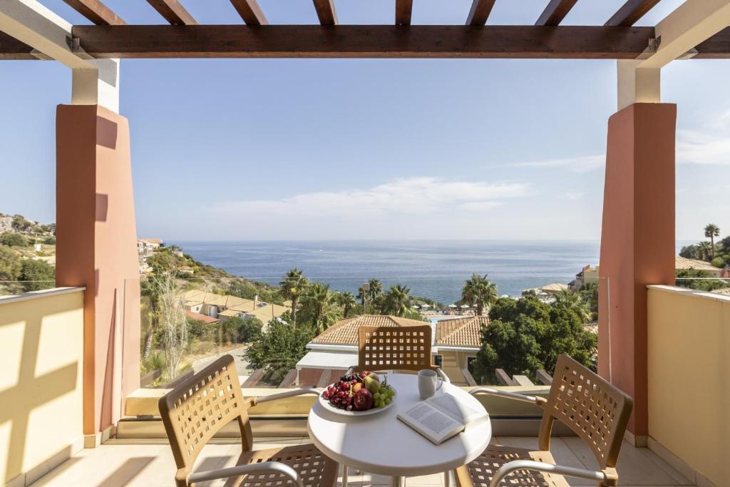 Skats no balkona uz jūru, kur redzams galds ar augļiem un grāmatu, apkārt zaļumi un mājas. Ideāla vieta atpūtai un relaksācijai.