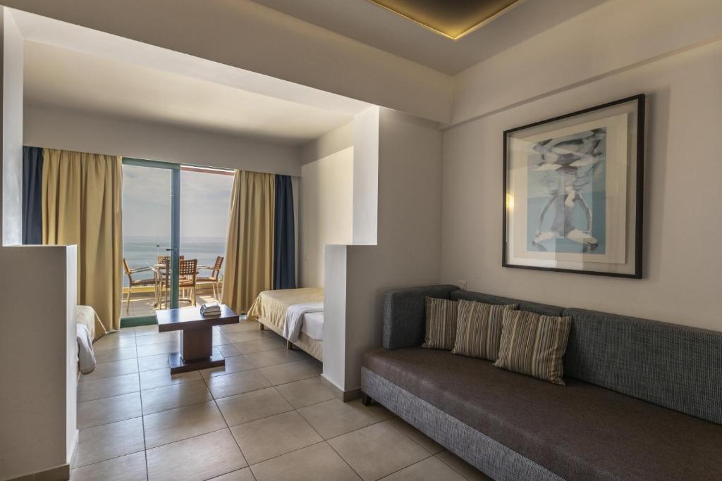 Moderns viesnīcas numurs ar skatu uz jūru, kurā ir divas gultas, dīvāns un galds. Labi apgaismots, ar plašām logu žalūzijām un mākslas darbu pie sienas.