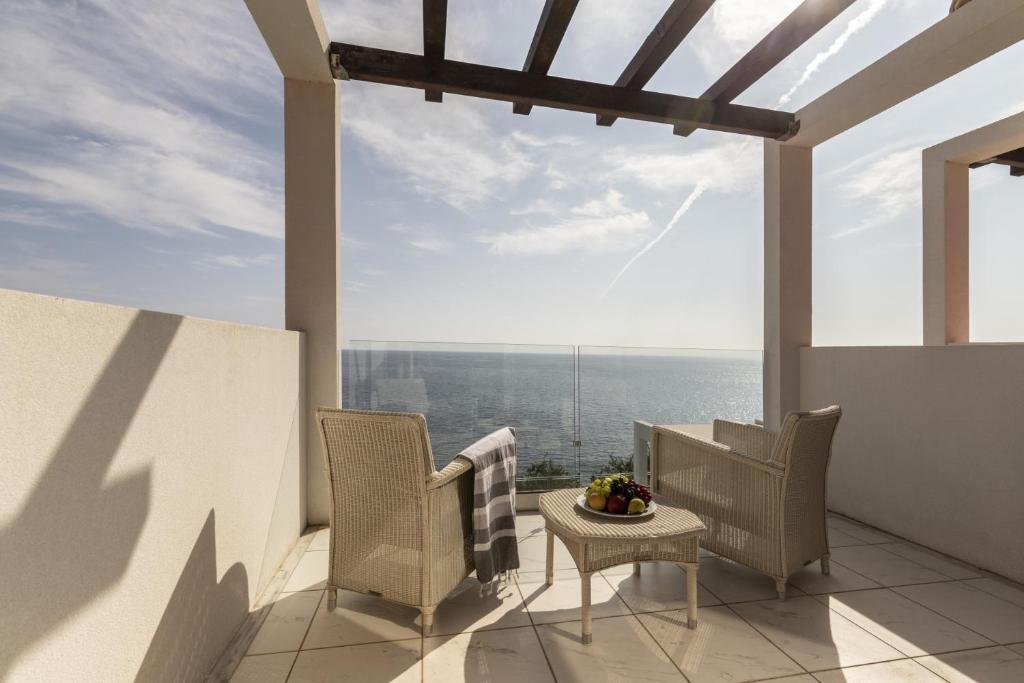 Terase ar skatu uz jūru, kur divas ērtas krēsli un galds ar augļiem rada relaksējošu atmosfēru. Saules gaisma un mīkstie mākoņi papildina mierīgu vidi.