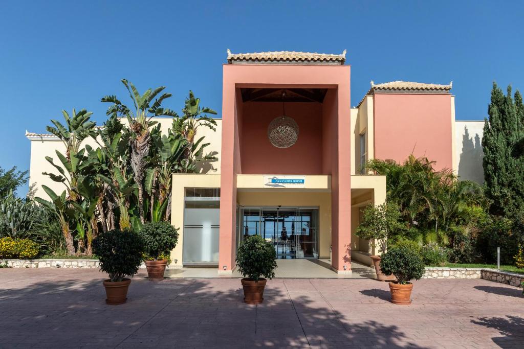 Mūsdienīga viesnīcas ieeja ar plašu terasi, dekorēta ar zaļiem augiem un palmām, sniedzot patīkamu atmosfēru.