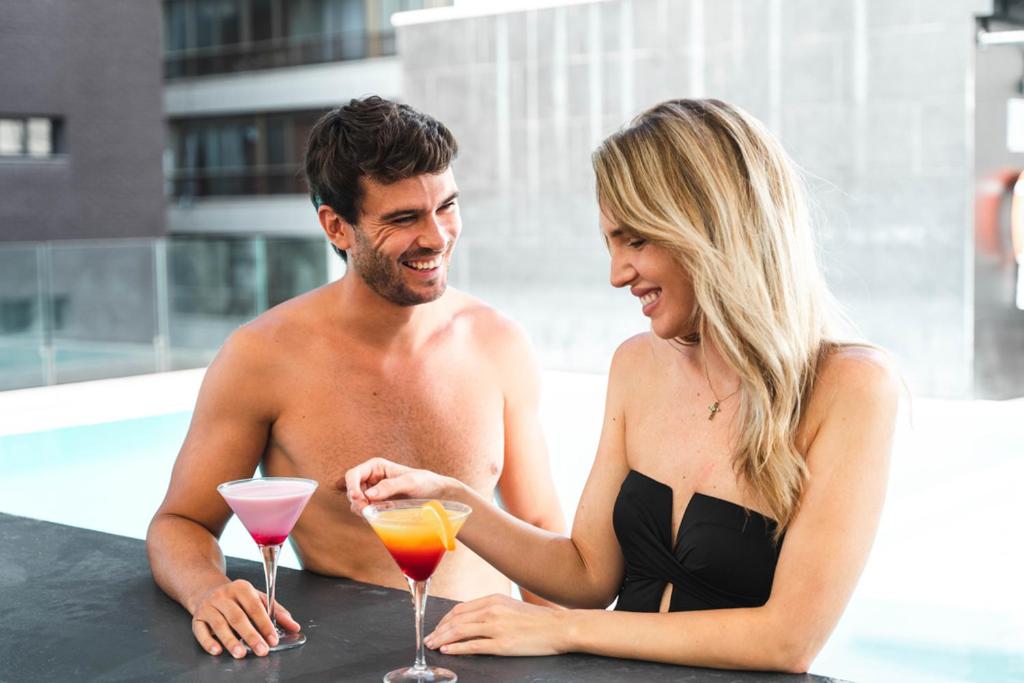 Jauns pāris bauda kokteiļus pie baseina, smaidot un izskatoties laimīgi. Fons ir moderns ēku komplekss.