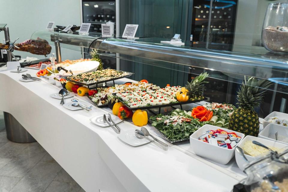 Bagātīgs bufetes galds ar dažādiem salātiem, augļiem un uzkodām, kas izkārtoti dekoratīvās traukos. Galds ir izveidots modernā stilā ar sudraba un baltas krāsas akcentiem.