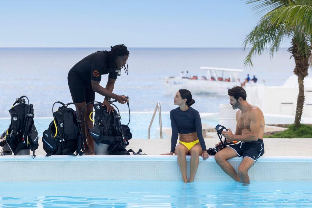 Scuba diving instrukcijas pie baseina, kur divi dalībnieki sarunājas ar instruktori. Fonā redzama jūra un laiva.