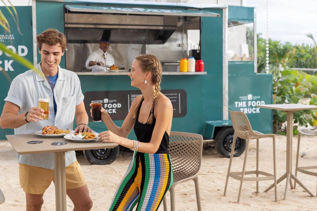 Jauna pārtikas auto apmeklējums, kurā jaunais pāris izbauda dzērienus un ēdienu, izskatās laimīgi un relaksēti. Fons ir pludmales vide ar zaļumiem un ērtām sēdvietām.
