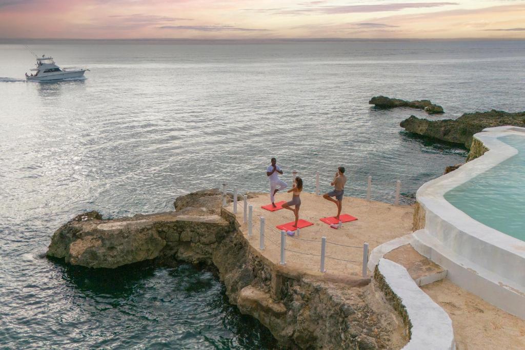 Meditācijas un jogas sesija pie jūras, ar trīs cilvēkiem, kas praktizē uz sarkanām paklājiem. Fons ir mierīgs okeāns un kuģis, kas peld pa ūdeni.