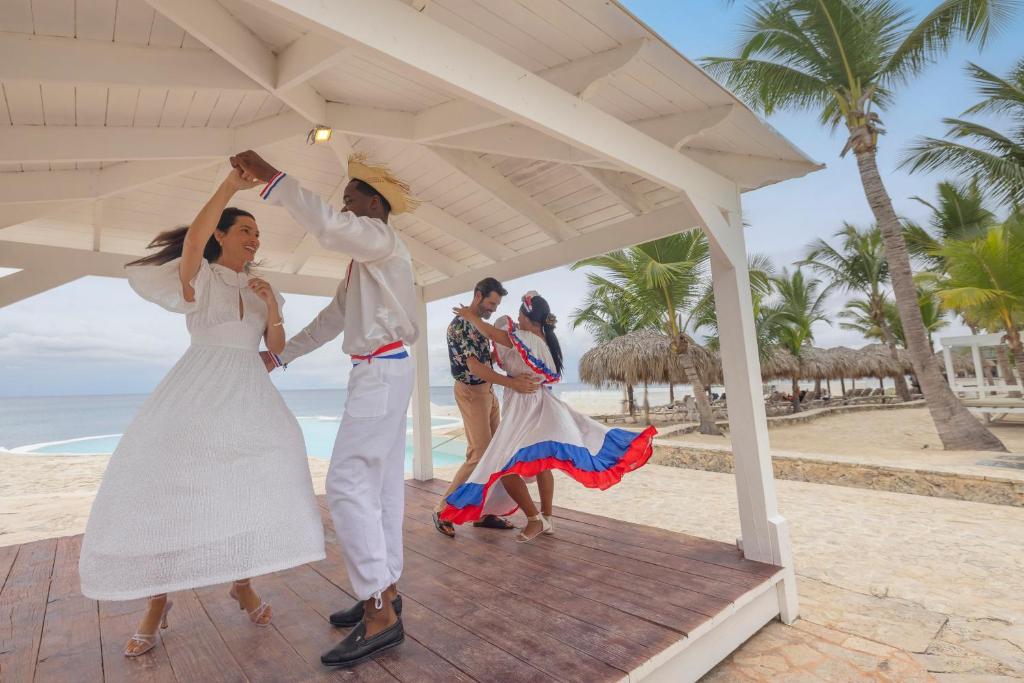Dejotāji tradicionālās tērpos izbauda deju pie pludmales paviljona, apkārt palmām un smiltīm.