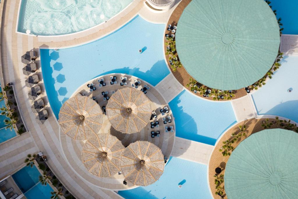 Viedoklis no augšas uz baseinu ar saulessargiem un atpūtas krēsliem, kas izvietoti ap ūdenstilpni, radot relaksējošu atmosfēru. Baseins ir iekrāsots zilos toņos ar palmām ap to.