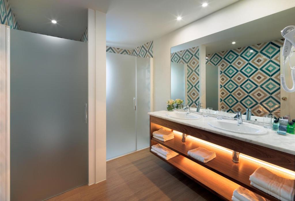 Mūsdienīgs vannas istabas interjers ar divām izlietnēm, stikla dušas kabīnēm un dekoratīvu sienu apdari. Vannas istabā ir koka skapīši un mīksti dvieļi.