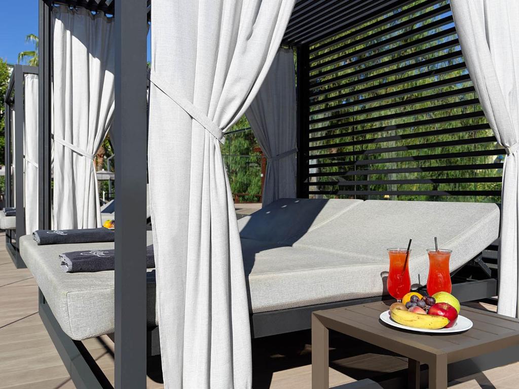 Mūsdienīgs atpūtas stūris ar gultu un baltām aizkariem, blakus atspirdzinoši kokteiļi un augļu šķīvis. Ideāla vieta relaksācijai un atpūtai pie baseina.