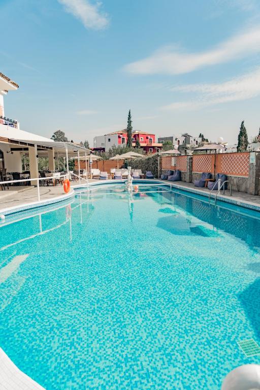 Peldbaseins ar skaidru zilu ūdeni un apkārtējo ainavu, kas ietver ērtus atpūtas krēslus un viesnīcas ēku fonā. Ideāla vieta relaksācijai un atpūtai saulainā dienā.