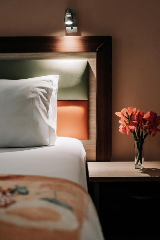 Mūsdienīgs guļamistabas interjers ar spilgtām krāsām, balto gultas veļu un ziedu kompozīciju uz naktsgaldiņa.