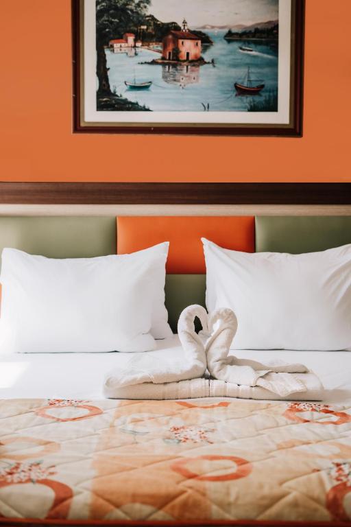 Gultas noformējums ar izrotātām dvielīšu gulbja figūrām un spilveniem, fona oranžā krāsā. Sienas māksla attēlo mierīgu skatu uz ūdeni ar laivām un ainavu.