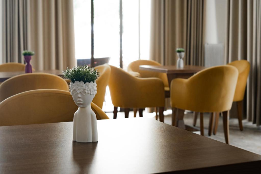 Mūsdienīgs interjers ar dzeltenām ērtām krēslām un dekoratīvām puķu podiem uz galda. Galvenais akcents ir stilizēts puķu pods cilvēka formā ar zaļām augu daļām.