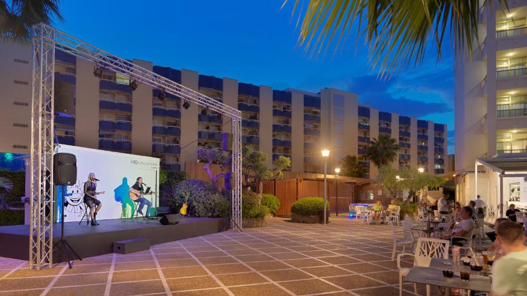 Āra koncerts ar izpildītājiem uz skatuves, apgaismots vakara laiks un viesi atpūšas tuvumā. Vides elementi, piemēram, augi un ēkas, veido patīkamu atmosfēru.