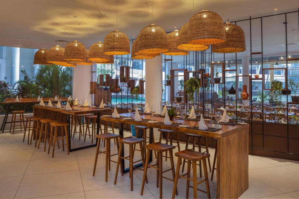 Mūsdienīgs restorāns ar koka mēbelēm un rotājošiem groza lampām, kas rada siltu atmosfēru. Telpā ir zaļumi un eleganta galda klāšana, kas nodrošina patīkamu ēdināšanas pieredzi.