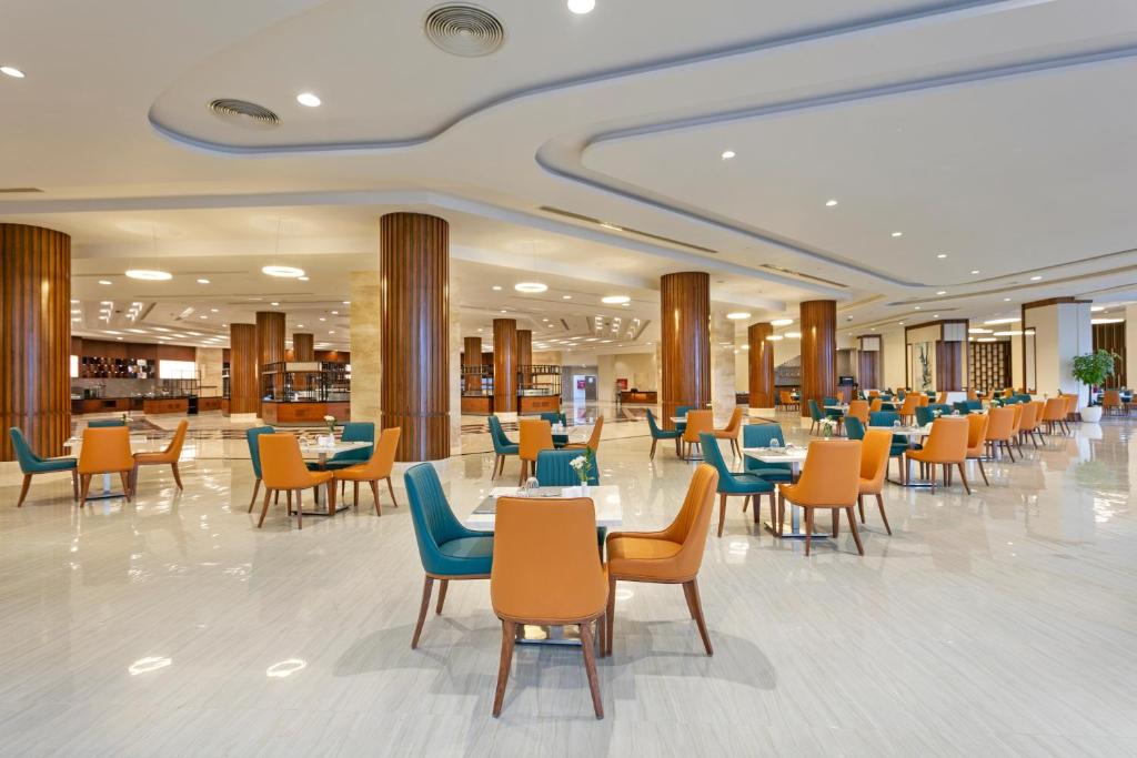 Mūsdienīgs restorāns ar plašu telpu, kas aprīkots ar krāsainām krēslām un elegantiem galdiem, radot patīkamu atmosfēru. Gaismas un dizaina elementi piešķir vietai mūsdienīgu noskaņu.