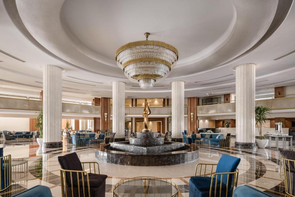 Mūsdienīga viesnīcas halle ar elegantu kristāla lustru un greznu strūklaku centrā, apkārt komfortablas sēdvietas un lieliskas arhitektūras elementi.