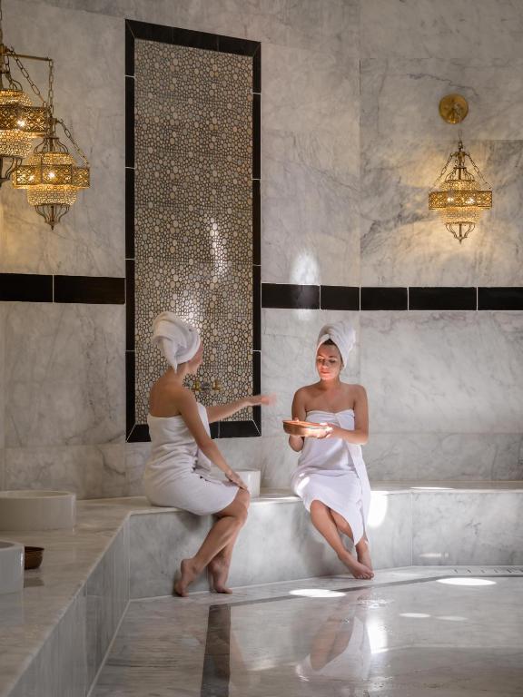 Divas sievietes, ietērptas dvielī, atpūšas greznā spa telpā ar marmora apdari un elegantām lampām. Viņas sarunājas un izbauda relaksējošu atmosfēru.