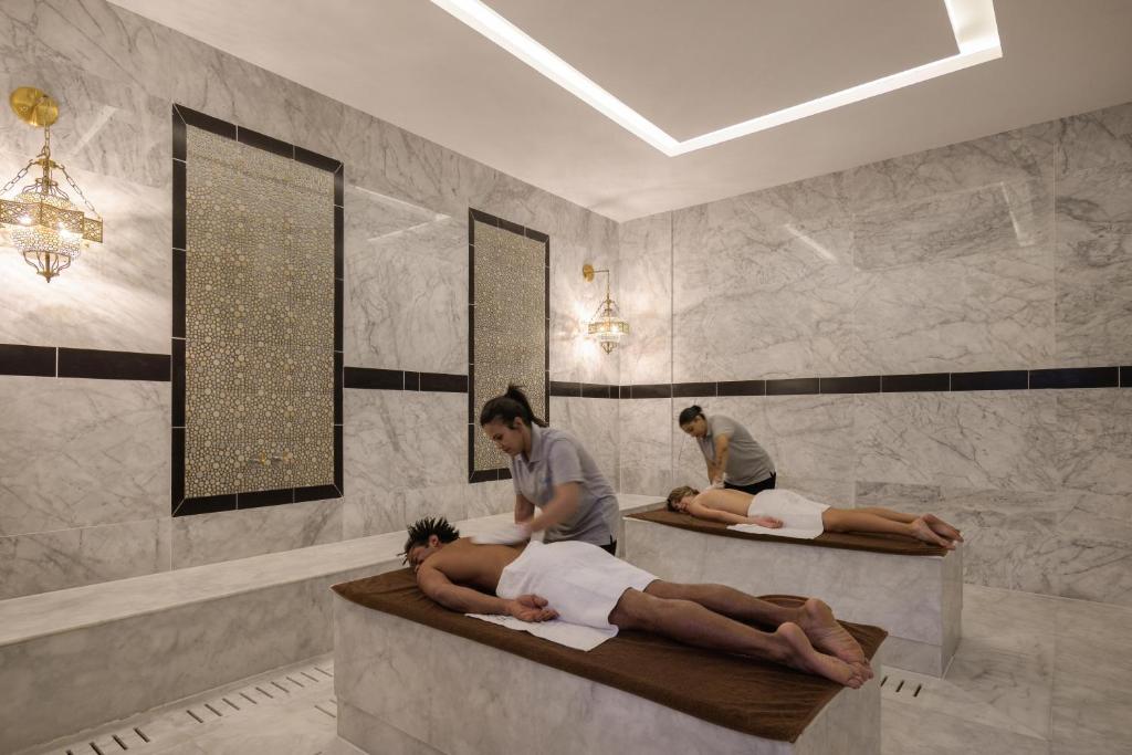 Spa telpa ar marmora sienām, kur divi masieri sniedz masāžas diviem klientiem uz masāžas galdiem. Gaisma ir maiga, radot relaksējošu atmosfēru.