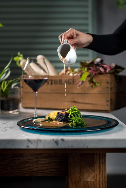 Garam masala mērce tiek pārlieta pār izsmalcinātu gaļas ēdienu, kas pasniegts uz elegantas plāksnes, blakus sarkanvīna glāze un dārzeņu rotājumi.