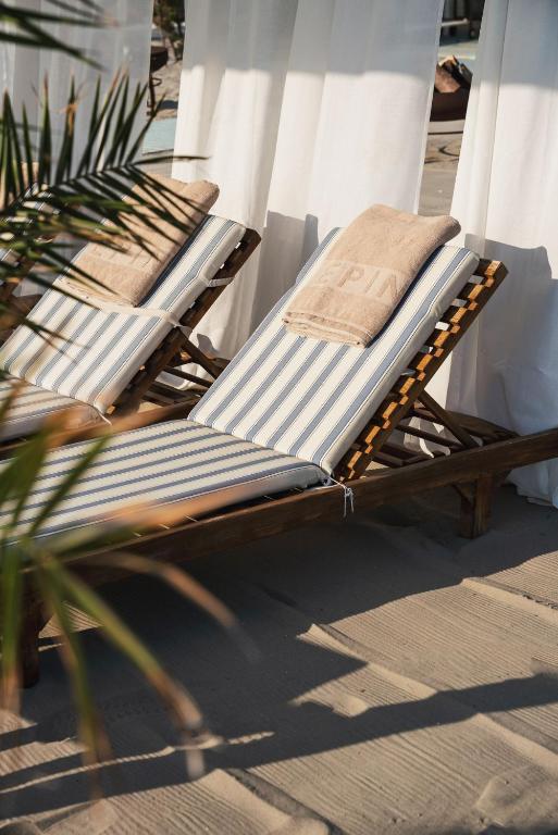 Atpūtas krēsli ar svītrām un dvieļiem, novietoti zem baltiem aizkariem pludmales vidē. Šis attēls rada relaksējošu atmosfēru.