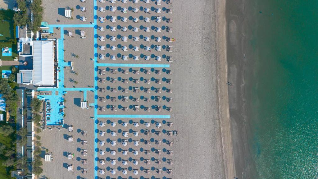 Pludmales skats no augšas, kur redzami rindā izvietoti sauļošanās krēsli un lietussargi, blakus modernai pludmales kafejnīcai. Jūra ar tīru, zilu ūdeni papildina relaksējošo atmosfēru.