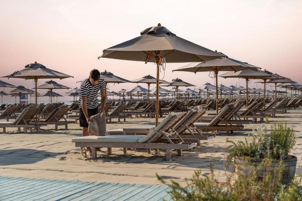 Pludmales ainava ar saulessargiem un atpūtas krēsliem, kurā vīrietis sakārto dvieli. Rīta gaisma un mierīga atmosfēra.