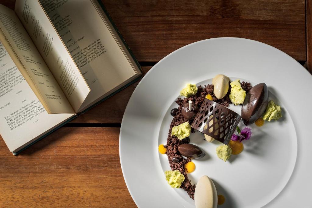 Grezna šokolādes deserts ar dažādām tekstūrām un krāsām, novietots uz baltas šķivja blakus atvērtai grāmatai.