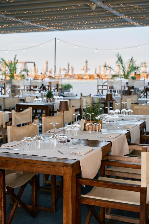 Restorāna iekārtojums ar koka galdiem un krēsliem, kas izkārtoti uz terases ar skatu uz ūdeni. Gaismas virtenes rada patīkamu atmosfēru.