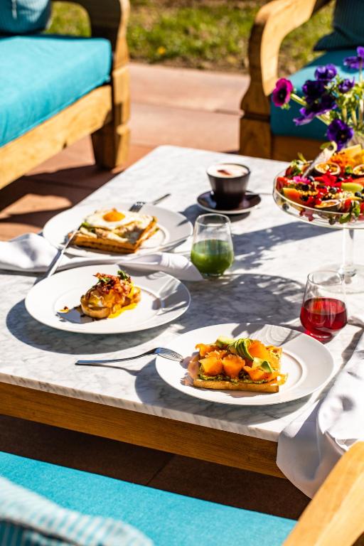 Skaista galda uzkodu izkārtojums, kurā redzami dažādi ēdieni, tostarp maizes šķēles ar augļiem un salātiem, kopā ar dzērieniem. Gaisīga un relaksējoša atmosfēra.