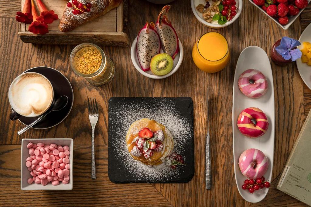 Svaigu brokastu galda kompozīcija ar pankūkām, augļiem, donātiem, kafiju un apelsīnu sulu, izkārtota uz koka galda.