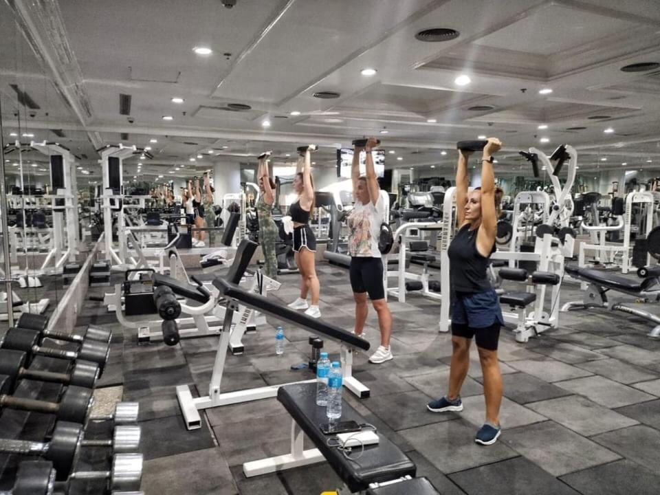 Trīs sievietes sporta zālē veic spēka treniņu ar hantelēm, paceļot tās virs galvas. Fons satur dažādas trenažieru iekārtas un spoguļus.