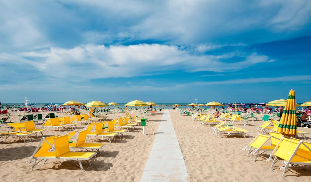 Pludmales ainava ar dzeltenām sauļošanās krēslām un lietussargiem, kas izkārtoti smilšainā pludmalē. Zilā debesis ar vieglām mākoņu joslām rada relaksējošu atmosfēru.