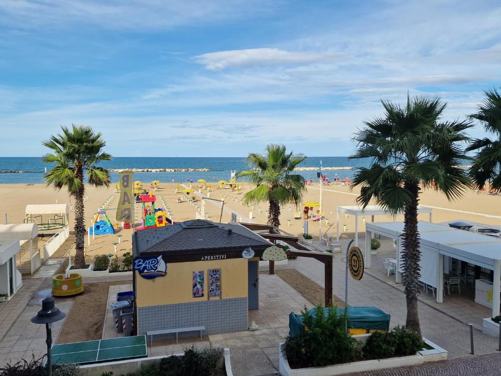 Pludmales ainava ar palmu kokiem un kafejnīcu, kas piedāvā uzkodas. Distantē redzama jūra un atpūtnieki pludmalē, kas izklāta ar sauļošanās krēsliem.