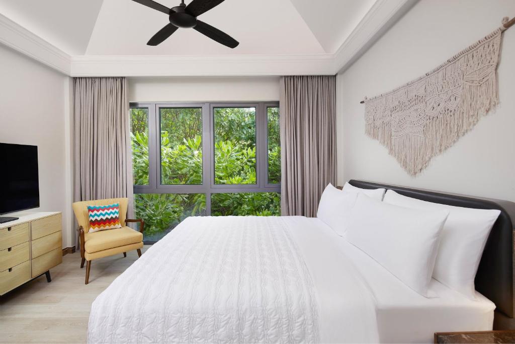 Mūsdienīgs guļamistabas interjers ar plašiem logiem, kas ļauj ieplūst dabiskajai gaismai. Gultā ir baltas gultas veļas, blakus ir ērta krēsla un televizora stends.