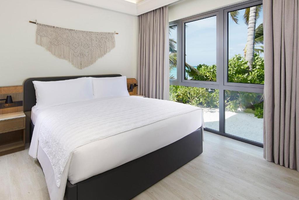 Mūsdienīga guļamistaba ar plašu logu, caur kuru redzams zaļš ainava un pludmale. Gultā ir balta gultasveļa un stilīgs interjers ar dabīgām krāsām.