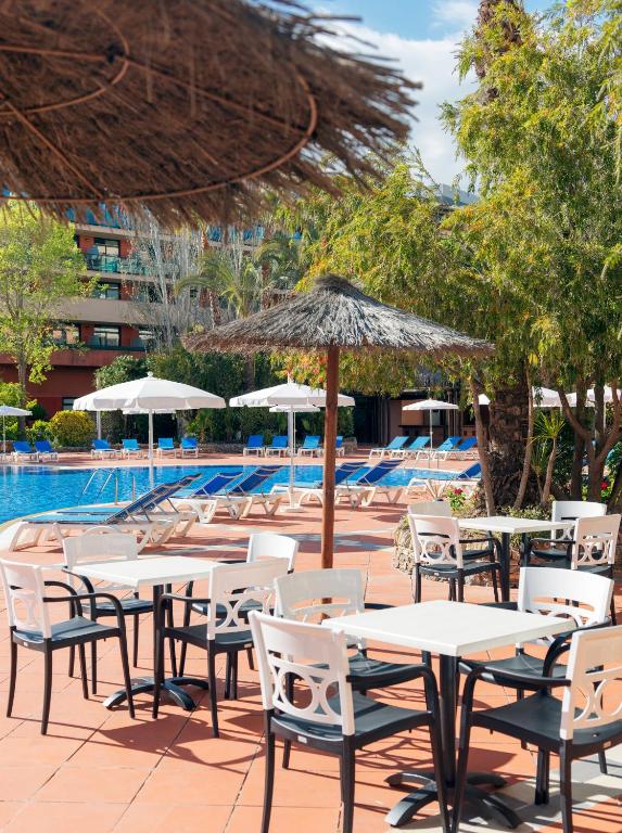 Peldbaseina zona ar sauļošanās krēsliem un ēdināšanas galdiem, apēnotiem ar lietus jumtiem, radot relaksējošu atmosfēru.