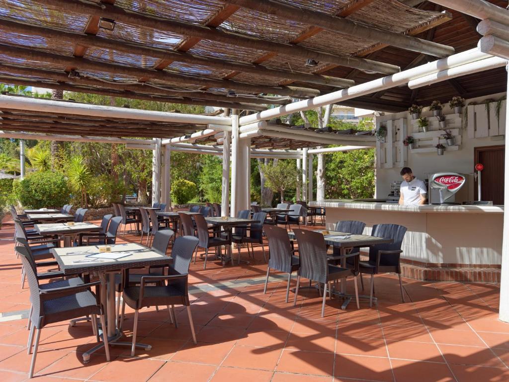 Ārtelpa restorāna terase ar galdiem un krēsliem, saulainā vidē, kur strādā viesmīlis pie bāra. Terase ir daļēji pārklāta ar koka konstrukciju, radot patīkamu atmosfēru.