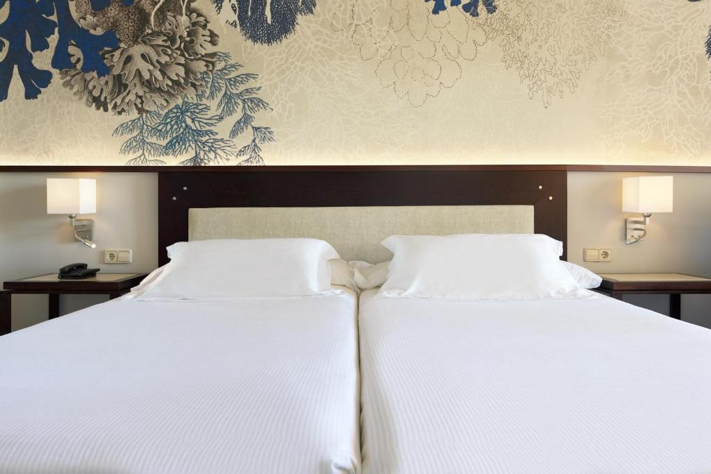 Mūsdienīgs viesnīcas numurs ar divām gultām, gaišām gultasveļām un dekoratīvu sienu ar jūras augu motīviem. Nummurā ir pieejama naktslampiņa un telefons.