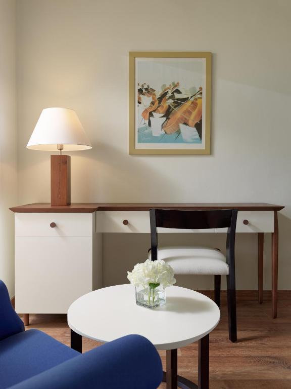 Mūsdienīgs interjers ar koka mēbelēm, lampu un gleznām, kas rada siltu un aicinošu atmosfēru. Uz galda ir ziedu kompozīcija.