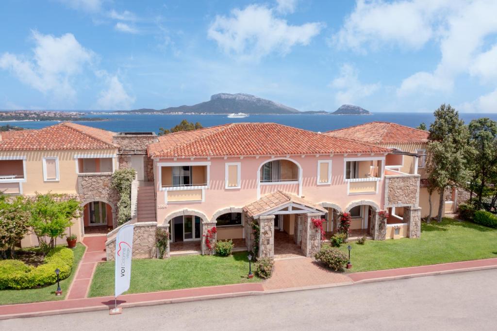 Elegants kūrorts ar rozā fasādi un akmens detaļām, kas atrodas pie jūras, ar skatu uz kalniem un zilo debesi. Apkārtne ir zaļa un labi kopta.