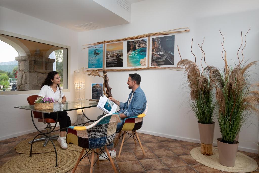 Gaišā un modernā telpā divi cilvēki sarunājas pie stikla galda, fonā redzami plakāti un dekoratīvi augi. Šī aina atspoguļo patīkamu un draudzīgu atmosfēru.