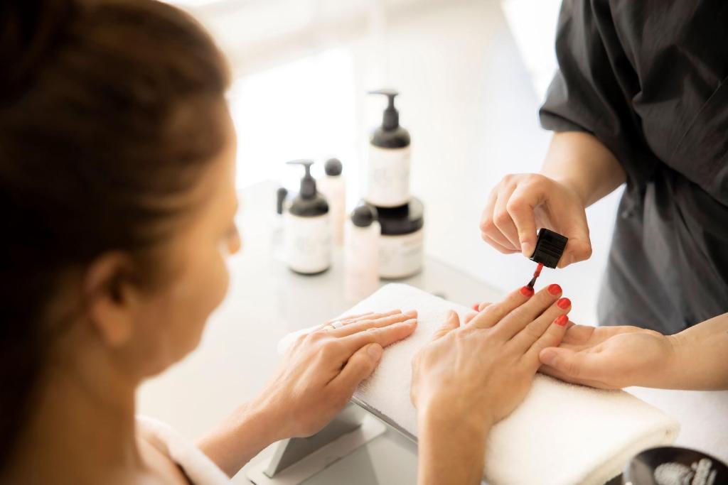 Nagu gleznu vizuālā māksla, manikīrs tiek veikts ar sarkano nagu laku, kamēr klients relaksējas. Fonā redzami kosmētikas produkti, kas piešķir greznu atmosfēru.